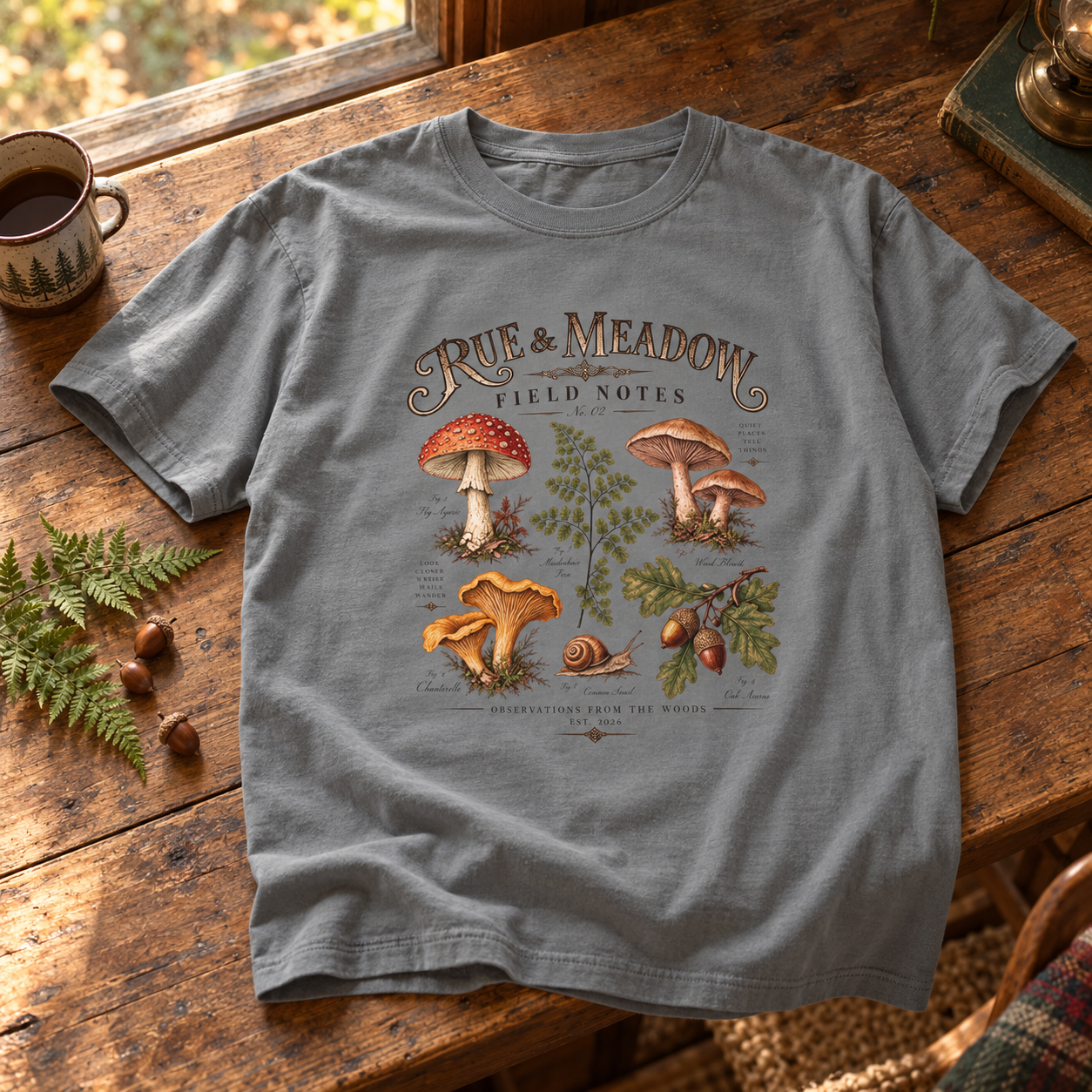 Gray Field Notes botanical T-shirt mockup with mushrooms, a fern, a snail, and oak leaves, styled on a rustic wooden table