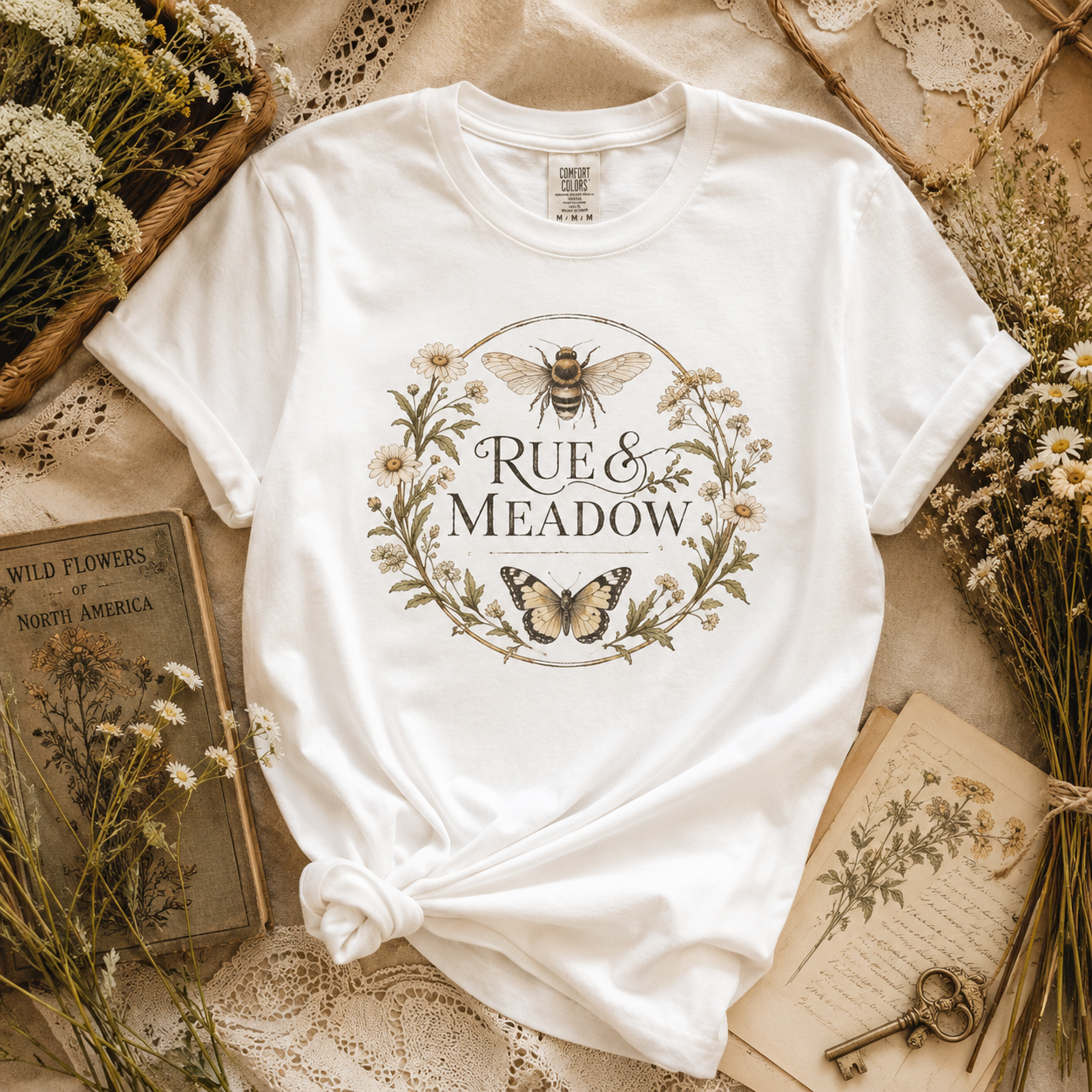 The original Rue and Meadow bee and butterfly floral wreath tee, styled on lace with wildflowers and antique botanical books
