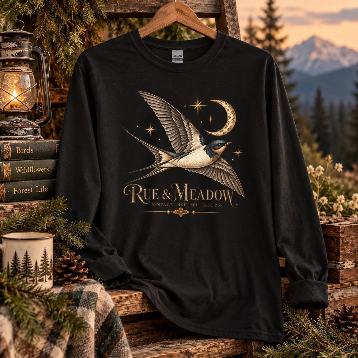 Black swallow long-sleeve tee with a crescent moon and stars, styled with a lantern, vintage books, and a mountain woodland view