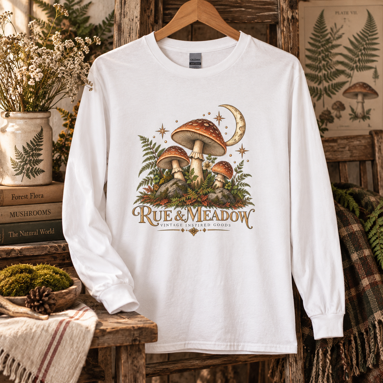 White mushroom and crescent moon long-sleeve tee on a wooden hanger, styled with ferns and vintage books